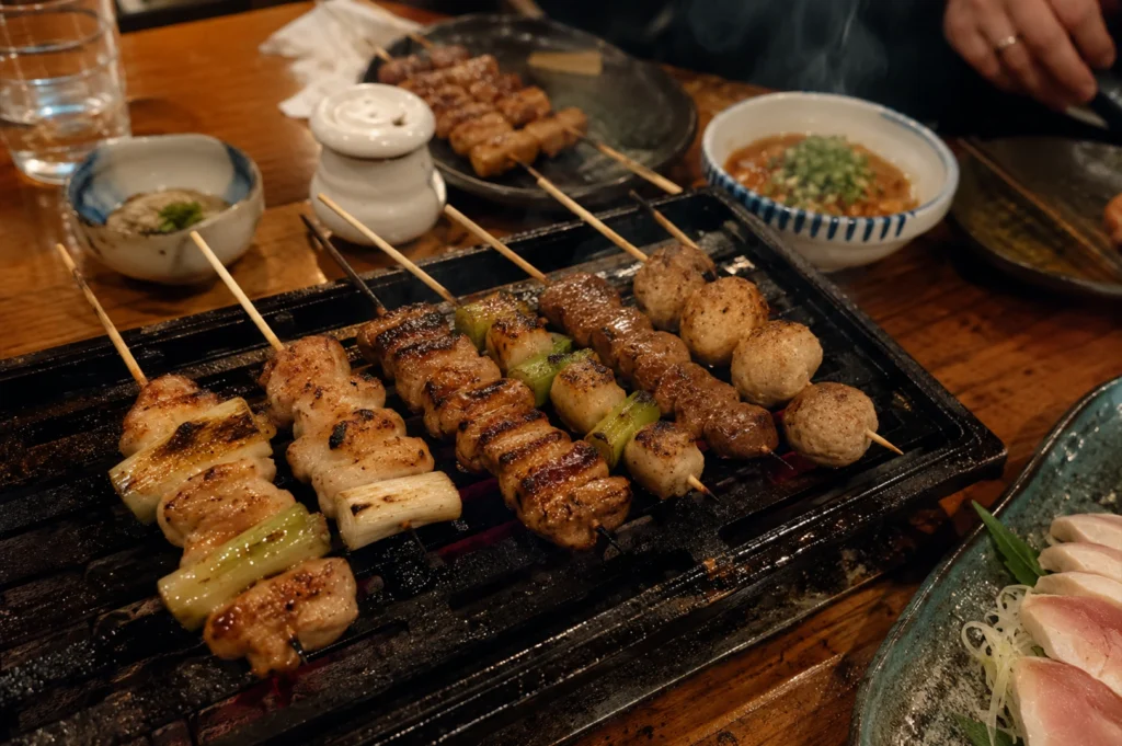 A top-down, angled shot displays a vibrant table setting inside an izakaya, centered around a small tabletop grill holding six freshly cooked yakitori skewers. From left to right, the skewers feature negima with charred green scallions, juicy chicken thigh, meat coated in a savory brown sauce, chicken skin, and round, lightly browned tsukune (chicken meatballs). Surrounding the grill on the warm wooden table are various side dishes, including a small blue-and-white bowl filled with a minced meat dish topped with chopped scallions, a plate of raw sliced chicken sashimi (toriwasa) in the bottom right corner, and a small white ceramic condiment shaker.