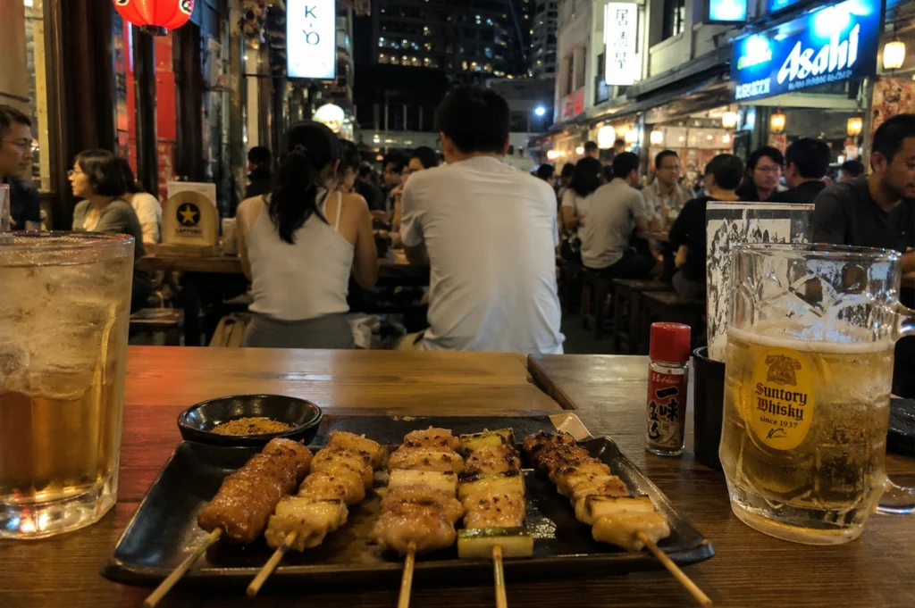 A vibrant evening scene from a diner's perspective at an outdoor eatery. In the foreground, a dark plate holds five yakitori skewers next to a tall glass of iced tea and a large mug of Suntory Whisky highball. The background is a bustling, blurred street scene at night with people sitting at wooden tables under bright neon signs and lanterns.