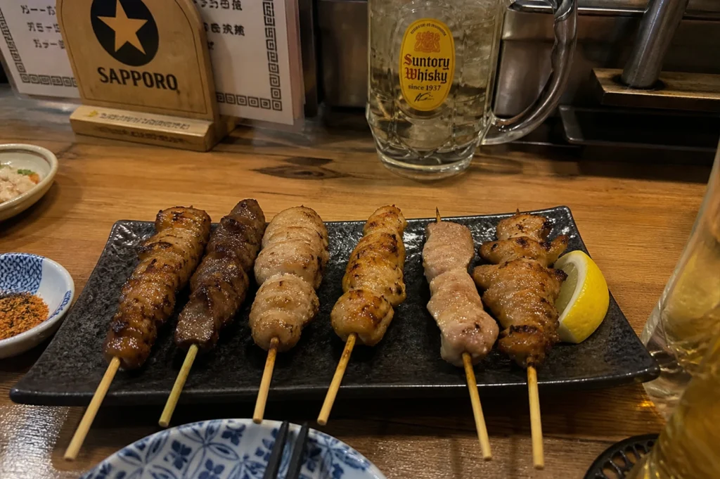A top-down view focused on a rectangular black plate featuring six assorted skewers, ranging from light-colored grilled chicken to darker, glazed meats, served with a fresh lemon wedge. Behind the plate, a large, dimpled glass mug of a Suntory Whisky highball filled with ice and lemon is prominent. In the background, a wooden menu stand with a Sapporo beer logo and small dishes of spices and appetizers are visible on the rustic wooden tabletop.