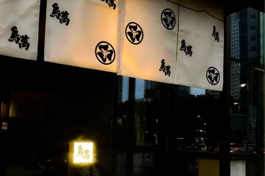White Japanese-style fabric curtains with black symbols and characters hang above a restaurant entrance at night. Warm lighting glows from inside the space, while reflections of nearby city buildings and street lights appear on the glass windows below.