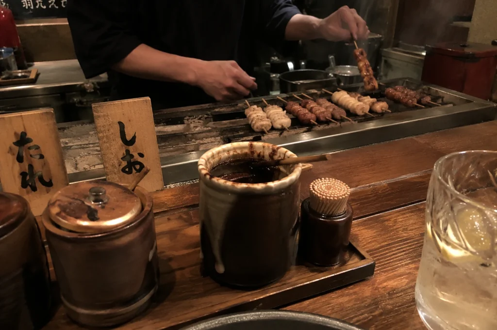 A warm, atmospheric shot of a yakitori prep station. In the foreground, rustic ceramic pots of "tare" (sauce) and a container of toothpicks sit on a wooden ledge. Behind the ledge, a chef is seen grilling several rows of skewers on a long, narrow charcoal grill. Wooden signs with Japanese calligraphy are propped up to the left.