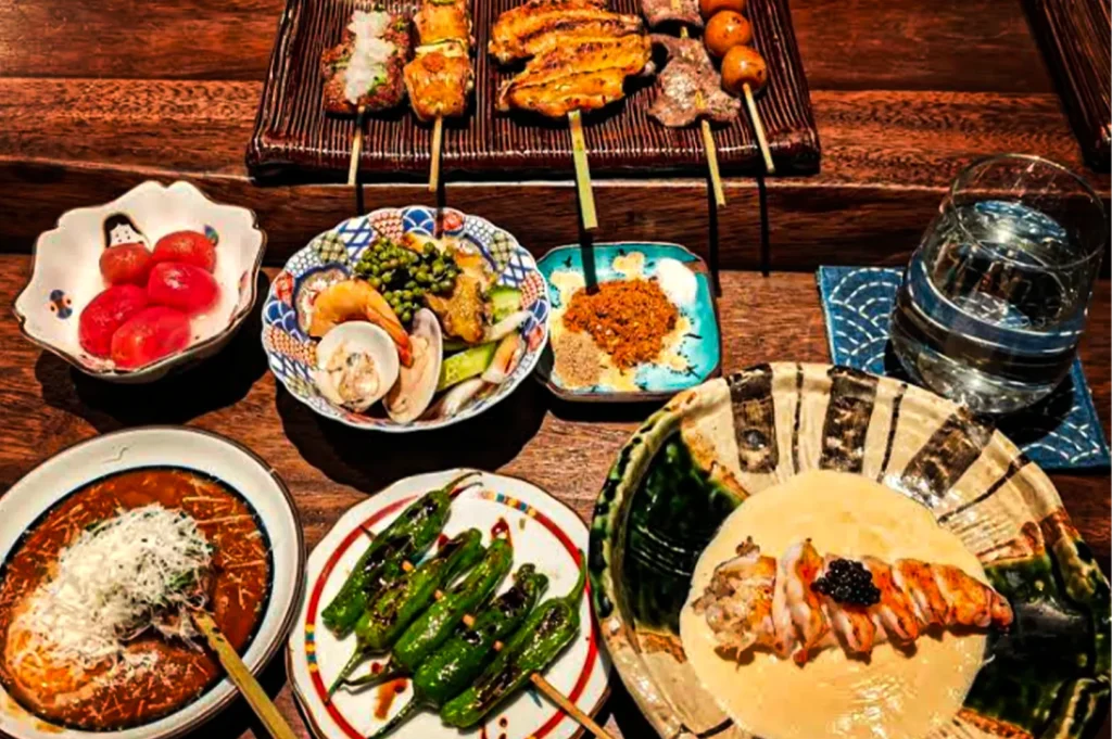 Overhead view of a Japanese-style meal spread on a wooden table featuring assorted grilled skewers, roasted peppers, seafood salad, cherry tomatoes, dipping sauces, and a bowl of creamy rice topped with sliced meat and roe. A glass of water and several colorful ceramic dishes complete the warm, cozy dining setup.