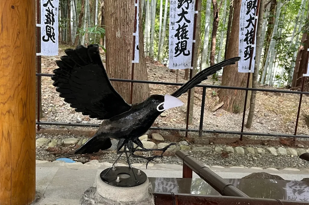 A black metal statue of the mythical three-legged crow Yatagarasu at a Japanese shrine, wearing a small white face mask, with bamboo trees in the background.