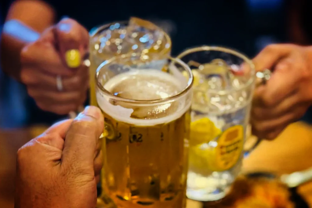 A close-up, first-person perspective shot of three people clinking glass mugs together in a celebratory toast over a table. In sharp focus in the foreground, a hand holds a glass mug filled with golden beer topped with thick white foam. Behind it, two other hands hold up highball glasses filled with iced drinks, one garnished with a lemon wedge featuring a yellow brand label on the glass. The background is softly blurred.