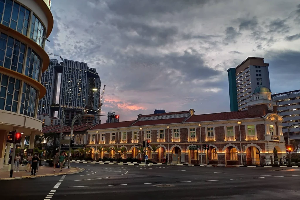 An architectural shot of a historic two-story brick building with white arched windows and trim, illuminated by warm yellow lights at dusk. The building sits on a street corner under a cloudy twilight sky. In the background, modern high-rise apartments and construction cranes provide a sharp contrast to the heritage architecture.
