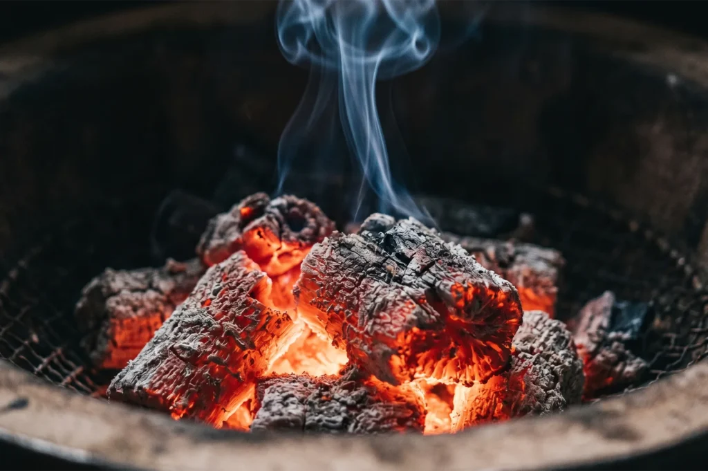 Close-up of glowing red hot binchotan charcoal embers with thin wisps of white smoke rising from a traditional Japanese grill.