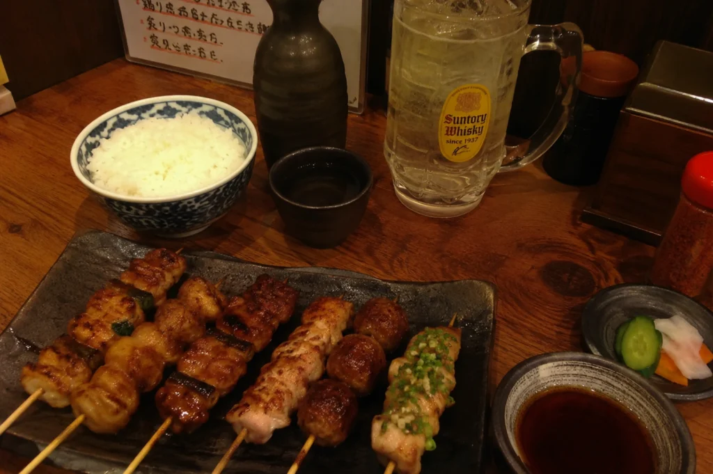 A high-angle, full table spread features a dark, rectangular ceramic plate lined with six assorted yakitori skewers, showcasing different textures from glistening glazed meats to rounded chicken meatballs (tsukune) and a skewer generously topped with finely chopped green scallions. The meal is arranged on a rich wooden table alongside traditional izakaya accompaniments: a blue-and-white patterned ceramic bowl filled with fluffy white rice, a small black cup paired with a dark ceramic sake carafe (tokkuri), a tall glass mug of a Suntory Whisky highball filled with ice, a small dish of dipping sauce, and a side of pickled cucumbers and daikon radish.