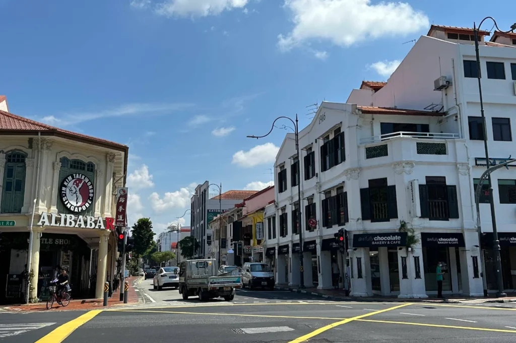 A bright, daytime street view of a Singaporean neighborhood featuring traditional shophouses. On the left, a building features a large circular sign for "Alibaba" and a Taekwondo school. On the right, a clean white building houses an "Awfully Chocolate" bakery and cafe. A white truck and several cars are positioned at the intersection under a clear blue sky.