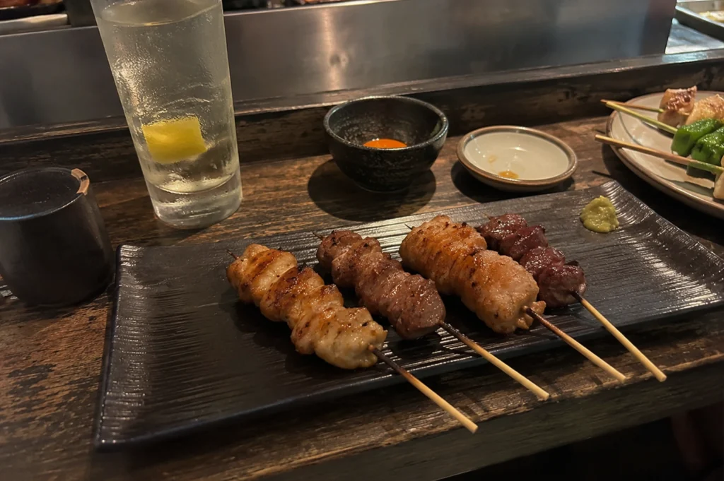 A high-angle, close-up shot of four yakitori skewers resting on a textured black rectangular plate. The skewers feature different cuts of grilled meat, varying from light golden-brown to a deep, charred reddish-brown. A small dollop of green wasabi sits on the corner of the plate. In the background, a tall glass of clear carbonated water with a lemon wedge is visible on a dark wooden bar counter, alongside a small black bowl containing an egg yolk and another empty dipping dish.