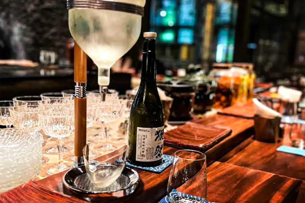 A sake-serving setup on a polished wooden counter featuring a chilled glass dispenser, crystal glasses, and a dark sake bottle with a Japanese label. Warm ambient lighting and blurred restaurant details in the background create an intimate bar atmosphere.