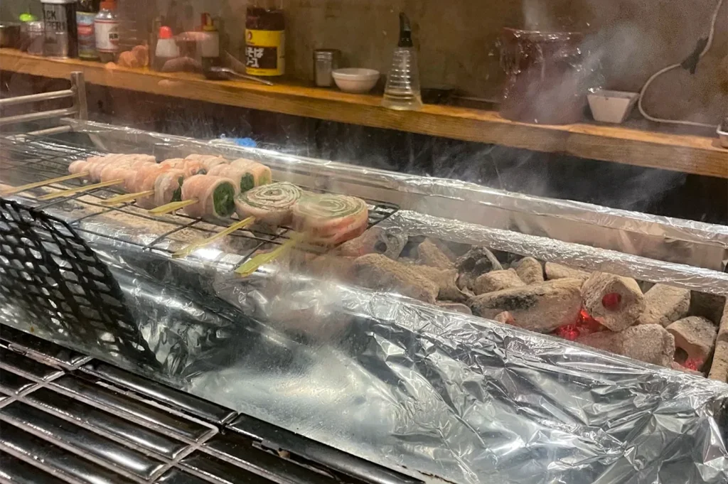 Meat and vegetable skewers, including pork-wrapped scallions, grilling over hot charcoal on a professional Japanese yakitori grill with visible smoke.