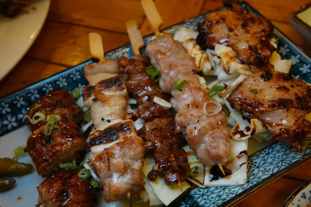 A close-up, top-down shot showcases a collection of cooked yakitori skewers arranged beautifully over a bed of crisp, raw white cabbage leaves on a blue-and-white patterned ceramic plate. The skewers feature a variety of textures and preparations, including heavily charred negima, deeply caramelized meats coated in a glossy, dark tare sauce, a lighter-colored skewer with a glistening sheen, and a larger piece of grilled chicken thigh on the right side. The dish is garnished with a scattering of finely chopped green onions, and a few small green chili peppers nestle in the bottom left corner of the plate.