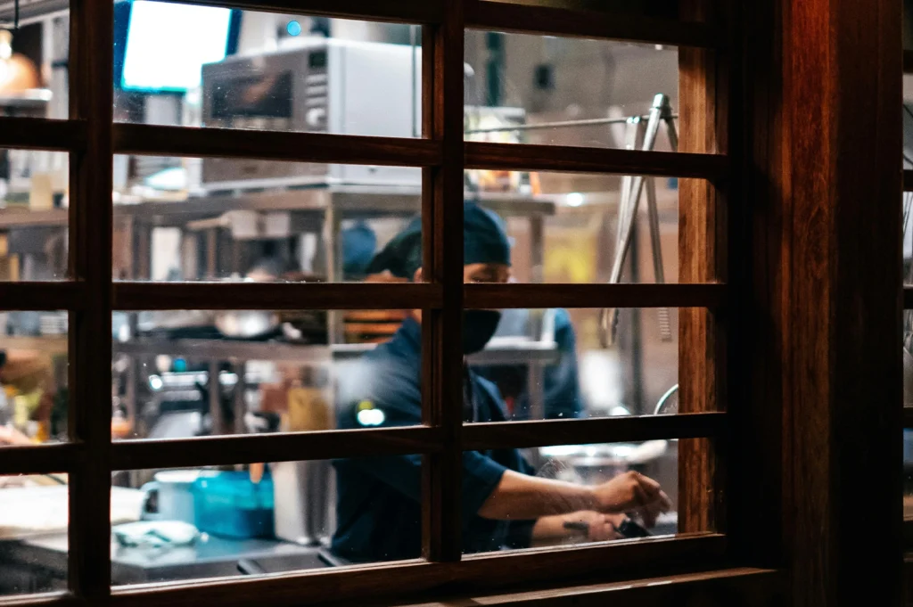 A candid, framed shot taken from outside the restaurant, looking through a dark wooden grid window. The focus is on a chef inside the kitchen, partially obscured by the wooden slats. He is wearing a dark uniform and a face mask, working near stainless steel kitchen equipment and a microwave. The lighting inside is bright and clinical compared to the warm tones of the dining area, offering a "behind-the-scenes" glimpse into the food preparation area.