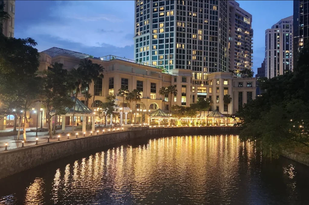 A serene night view of a riverfront promenade. Warm lights from a multi-story hotel and riverside restaurants reflect gold ripples onto the dark water. The architecture is a mix of classical columns and glass-domed gazebos, with lush green trees lining the walkway on the right.