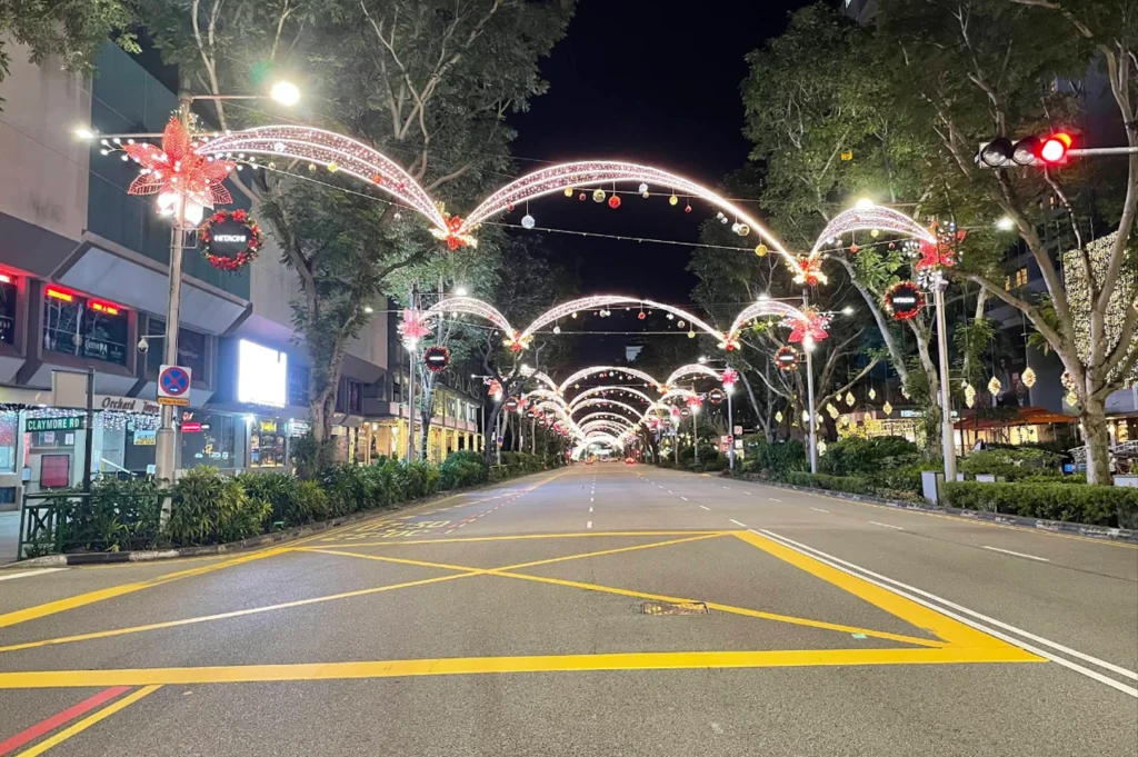 A wide-angle shot of Orchard Road in Singapore at night, decorated with elaborate Christmas light arches. The arches are adorned with red floral motifs and hanging ornaments, stretching over a wide, empty asphalt road marked with yellow lines. The sidewalks are lined with trees and the glowing storefronts of luxury shopping malls.