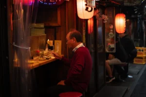 A profile, eye-level shot captures a man wearing a red sweater and glasses eating alone at an outdoor wooden counter of a narrow alleyway izakaya, representing the file "1-After the Rush Why Yakitori Becomes a Ritual.webp". He sits on a red stool, using chopsticks to pick at small dishes on the bar counter, which is partially partitioned by a translucent plastic curtain on the left. The scene is illuminated by the warm, atmospheric glow of traditional paper lanterns hanging overhead—one white and one red—casting a vivid red reflection on the glass doors behind him. In the background, other customers sit along the cramped alleyway stall, creating a quintessential, cozy nocturnal dining scene.