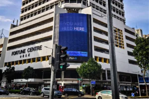A low-angle, medium-wide shot captures the distinctively tiered, white facade of the Fortune Centre building at a city street corner, representing the file "1-Best Fortune Centre Food for Japanese Dining.webp". The building features bold, black lettering reading "FORTUNE CENTRE" with matching Chinese characters underneath on its left wing, while a massive, vertical digital billboard dominates the central nook of the structure. The electronic display shows a deep blue ocean horizon beneath the words "YOU ARE HELD HERE" in a clean white font, with "Plan B" branded at the very bottom. In the foreground, a gray traffic light pole stands near an active intersection where several cars, including a white sedan and a black SUV, are driving or waiting, framed under a lightly clouded blue sky.