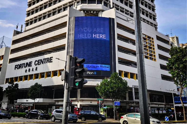 A low-angle, medium-wide shot captures the distinctively tiered, white facade of the Fortune Centre building at a city street corner, representing the file "1-Best Fortune Centre Food for Japanese Dining.webp". The building features bold, black lettering reading "FORTUNE CENTRE" with matching Chinese characters underneath on its left wing, while a massive, vertical digital billboard dominates the central nook of the structure. The electronic display shows a deep blue ocean horizon beneath the words "YOU ARE HELD HERE" in a clean white font, with "Plan B" branded at the very bottom. In the foreground, a gray traffic light pole stands near an active intersection where several cars, including a white sedan and a black SUV, are driving or waiting, framed under a lightly clouded blue sky.