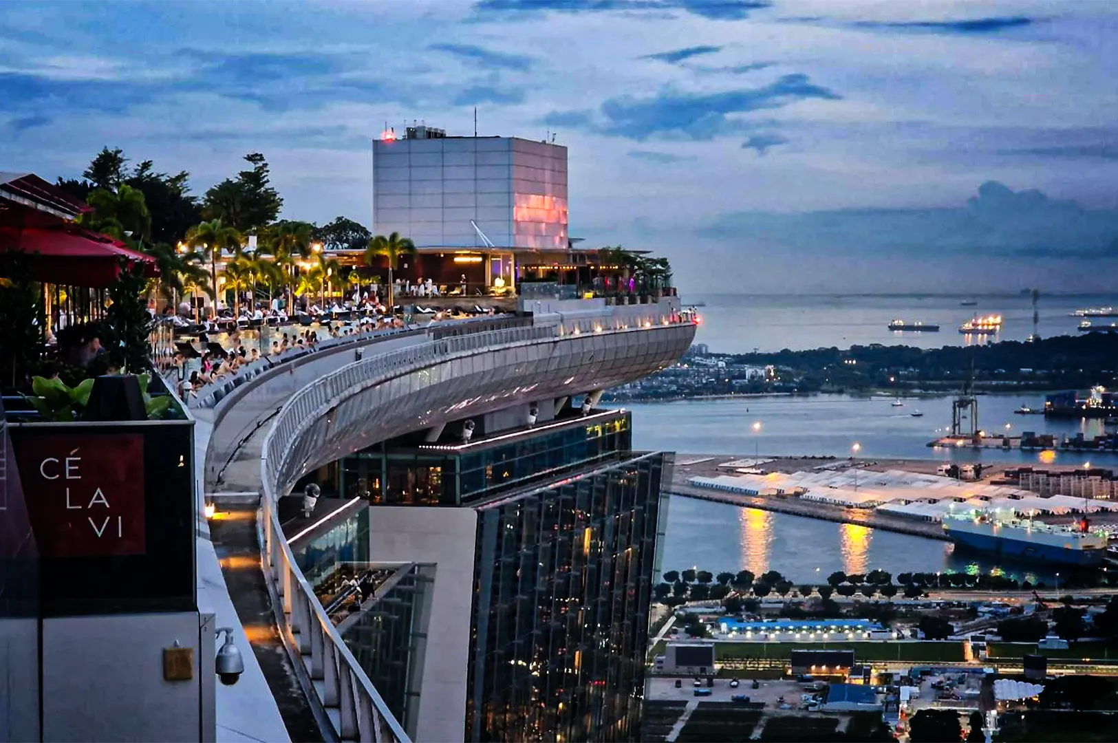 A high-angle, wide shot captures the expansive, curved sky deck of the Marina Bay Sands resort at dusk, representing the file "1-Best Yakitori in Marina Bay Sands Food 6 Spots.webp". On the left, the lively rooftop features the CÉ LA VI restaurant and a crowded infinity pool lined with palm trees and glowing lounge lights under a cloudy evening sky. The cantilevered observation deck juts out dramatically over the modern glass facade of the hotel tower below. In the background, a sprawling view of the Singapore Strait unfolds, dotted with large cargo ships and industrial docks illuminated by warm harbor lights, creating a sophisticated and vibrant urban landscape.