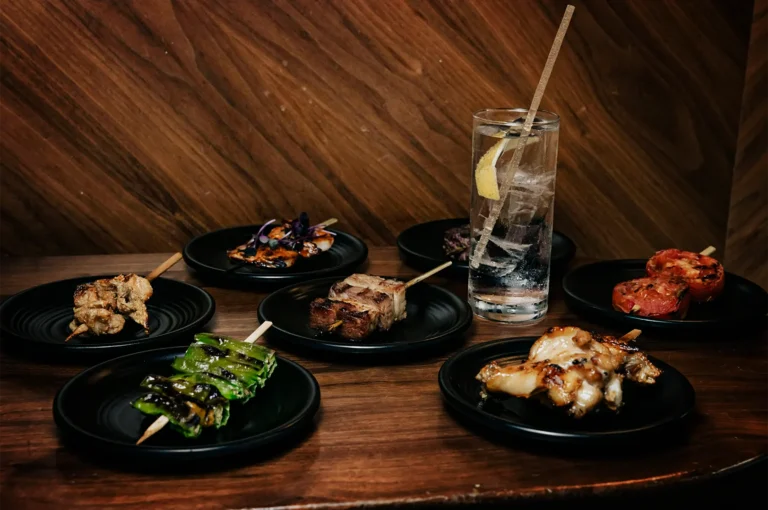 An eye-level shot of several small black plates arranged on a dark wooden table, each holding a different grilled Japanese skewer. The dishes include charred green peppers, cubed pork belly, grilled chicken, and seared tomatoes. A tall, clear highball glass filled with ice, a lemon wedge, and a clear fizzy drink stands among the plates against a warm, wood-paneled wall backdrop.