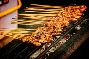 A high-angle close-up shot captures rows of Japanese chicken skewers cooking over an open flame, representing the file "1-Japanese Chicken Skewers Explained.webp". The yakitori skewers are tightly lined up across a narrow, dark metal grill grate, with their long wooden handles extending out to the left side of the frame. The chicken meat is deeply browned and glistening with a savory glaze, showing dark, crispy charred spots from the high heat. Wisps of smoke gently rise from the meat as it cooks over glowing red-hot charcoal embers visible beneath the grate, creating a warm, appetizing, and traditional street-food aesthetic.