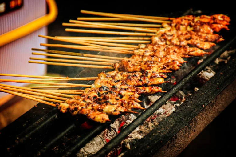 A high-angle close-up shot captures rows of Japanese chicken skewers cooking over an open flame, representing the file "1-Japanese Chicken Skewers Explained.webp". The yakitori skewers are tightly lined up across a narrow, dark metal grill grate, with their long wooden handles extending out to the left side of the frame. The chicken meat is deeply browned and glistening with a savory glaze, showing dark, crispy charred spots from the high heat. Wisps of smoke gently rise from the meat as it cooks over glowing red-hot charcoal embers visible beneath the grate, creating a warm, appetizing, and traditional street-food aesthetic.