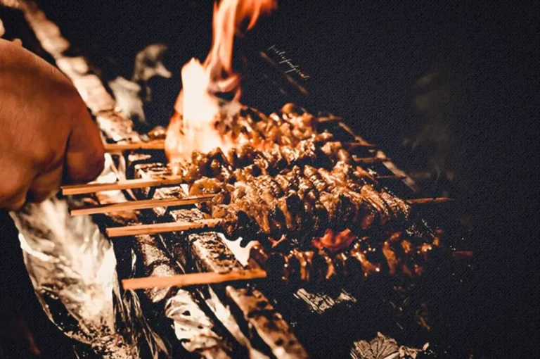 A dramatic, close-up shot captures a row of yakitori skewers cooking over an intense, open-flame grill, representing the file "1-Mastering the Grill Reflections from Kai Yakitori.webp". Set against a stark black background, a bright orange flame erupts from the hot charcoal below, licking up through the metal grates and searing the closely packed, glistening meat skewers. On the left, a chef's hand is visible in partial profile, deftly managing the wooden ends of the skewers. The high-contrast, moody lighting accentuates the textured, textured canvas-like grain of the image, the crinkled aluminum foil lining the edge of the grill, and the sizzling, smoky atmosphere of an authentic Japanese kitchen.
