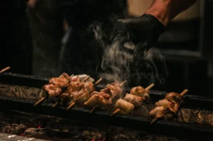 A close-up, eye-level shot captures a row of yakitori skewers cooking over an open-fire grill, representing the file "1-Not Just a Stick The Role of Bamboo.webp". Several bamboo skewers threaded with pieces of chicken are neatly aligned across the narrow metal bars of a traditional grill, with glowing red embers visible below them. Wisps of white smoke rise from the sizzling meat, creating a dynamic sense of heat and aroma. On the right, a chef's hand clad in a black nitrile glove hovers over the food, preparing to turn the skewers against a dim, darkly shadowed background.