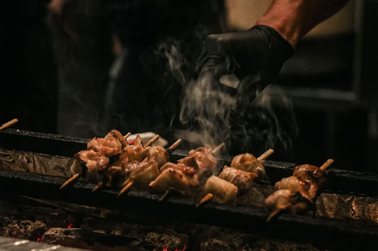 A close-up, eye-level shot captures a row of yakitori skewers cooking over an open-fire grill, representing the file "1-Not Just a Stick The Role of Bamboo.webp". Several bamboo skewers threaded with pieces of chicken are neatly aligned across the narrow metal bars of a traditional grill, with glowing red embers visible below them. Wisps of white smoke rise from the sizzling meat, creating a dynamic sense of heat and aroma. On the right, a chef's hand clad in a black nitrile glove hovers over the food, preparing to turn the skewers against a dim, darkly shadowed background.