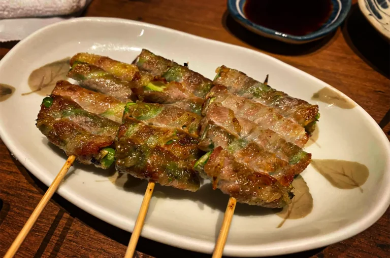 A close-up, slightly high-angle view of three wooden skewers holding grilled pork belly rolls wrapped around green asparagus or scallions, served on an oval white ceramic plate with a subtle leaf pattern. The meat is beautifully seared and lightly charred, resting on a dark wooden table next to a small blue-rimmed dipping dish filled with dark soy sauce in the background.
