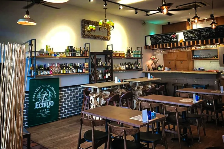 An eye-level, medium shot captures the warm, inviting interior of a Japanese bar and restaurant, representing the file "1-TORIMARO Singapore Review.webp". The dining area features a mix of dark and light wood tables paired with matching wooden chairs, organized across a polished hardwood floor. Along the left wall, a partially brick-textured lower section sits beneath extensive wall shelving heavily stocked with various sake and liquor bottles, flanked by a green banner reading "Echigo, Japan's First Micro-Brewery." In the background on the right, a wooden bar counter with high stools stands under hanging wine glasses and a dark chalkboard menu, while a blend of modern track lighting, industrial pendant lamps, and a colorful stained-glass chandelier casts a cozy, ambient glow over the entire space.
