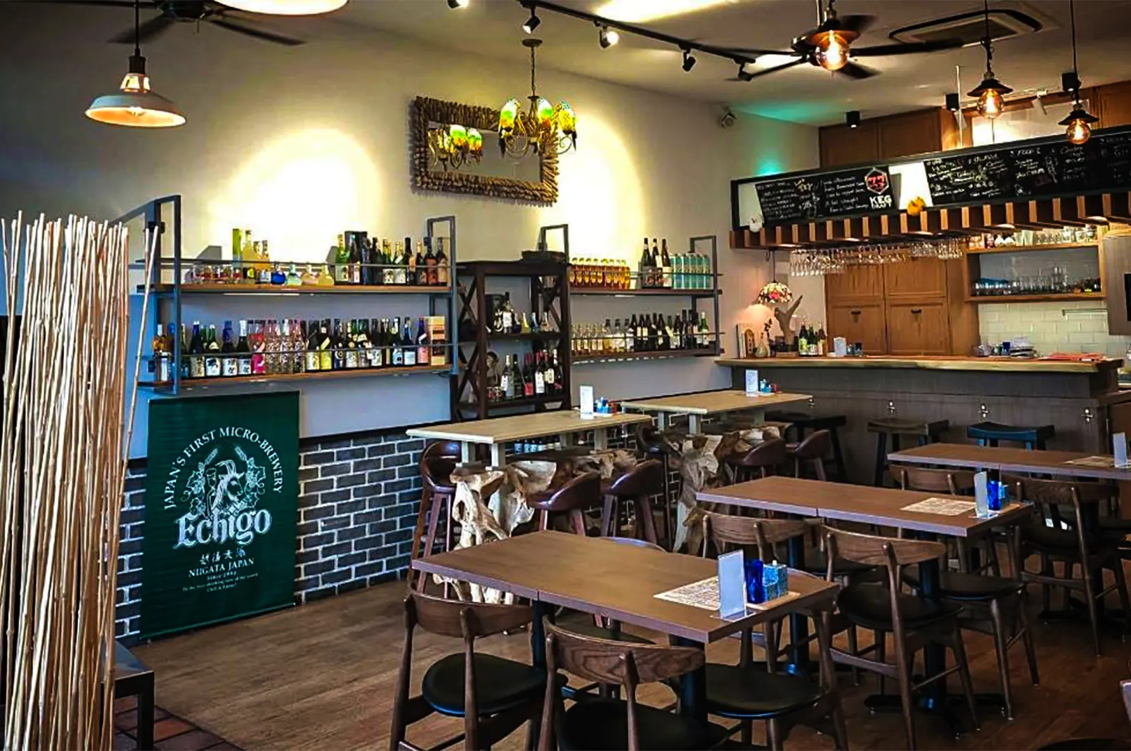 An eye-level, medium shot captures the warm, inviting interior of a Japanese bar and restaurant, representing the file "1-TORIMARO Singapore Review.webp". The dining area features a mix of dark and light wood tables paired with matching wooden chairs, organized across a polished hardwood floor. Along the left wall, a partially brick-textured lower section sits beneath extensive wall shelving heavily stocked with various sake and liquor bottles, flanked by a green banner reading "Echigo, Japan's First Micro-Brewery." In the background on the right, a wooden bar counter with high stools stands under hanging wine glasses and a dark chalkboard menu, while a blend of modern track lighting, industrial pendant lamps, and a colorful stained-glass chandelier casts a cozy, ambient glow over the entire space.