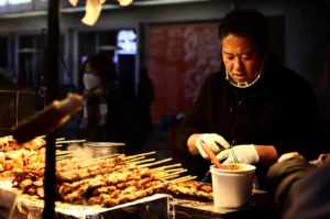 A medium, eye-level shot captures a vendor focused on preparing food at an outdoor street stall at night, representing the file "1-Taste of Anticipation Why the First Skewer Feels Different.webp". The man, wearing a black jacket, a clear plastic chin shield, and white work gloves, stands behind his counter working over a small container with a red brush tool. In the foreground, large stacks of grilled meat skewers sit on a metal tray, steaming slightly under the warm stall illumination. The background is softly blurred, showing glimpses of a dark city street, a pedestrian wearing a face mask and scarf, and the faint glow of nighttime storefronts.