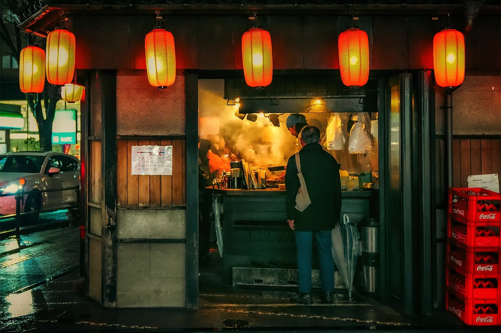 A customer standing at the entrance of a small Japanese street food restaurant at night, with glowing red lanterns hanging above the storefront, steam rising from the open kitchen, chefs cooking behind the counter, and wet city streets reflecting lights outside.