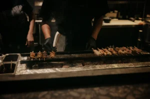 A close-up shot of a chef in a dimly lit commercial kitchen cooking meat skewers on a long, narrow charcoal grill. The chef, wearing black gloves, uses one hand to carefully turn a row of yakitori skewers on the left, while another long line of skewers cooks over glowing hot coals on the right. Wisps of smoke rise from the grill, and stainless steel kitchen counters are visible in the background.