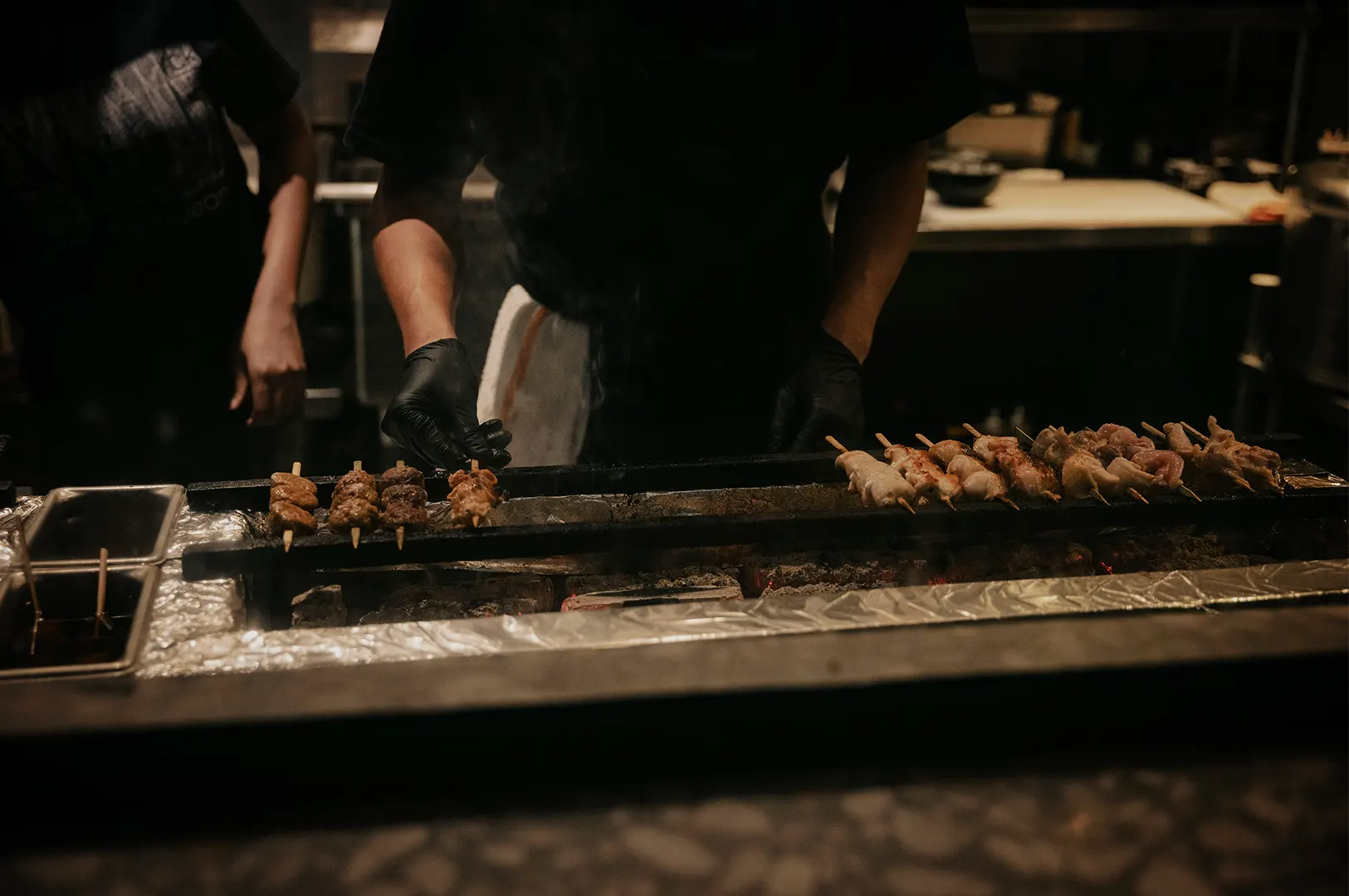 A close-up shot of a chef in a dimly lit commercial kitchen cooking meat skewers on a long, narrow charcoal grill. The chef, wearing black gloves, uses one hand to carefully turn a row of yakitori skewers on the left, while another long line of skewers cooks over glowing hot coals on the right. Wisps of smoke rise from the grill, and stainless steel kitchen counters are visible in the background.