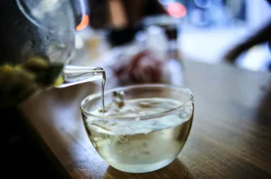 An eye-level shot of a professional bartender or chef in a white chef's coat at a clean, light wood workstation inside a modern restaurant. The chef is looking down, using a long knife to slice a piece of fish on a large rectangular cutting board. To the left, several small blue-and-white checkered condiment bowls are neatly arranged in a row. The background features a large glass window looking out at a dark garden at night, with a striking display of several Japanese chef knives arranged vertically on a stand on the right.