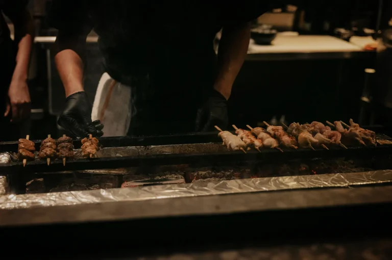 An eye-level, close-up shot captures a chef meticulously cooking Japanese yakitori in a dimly lit restaurant kitchen, representing the file "1-The Things You Notice.webp". Wearing a black uniform and black latex gloves, the chef's hands are focused on a long, narrow charcoal grill lined with aluminum foil, where glowing hot coals emit a light stream of smoke. Arrayed across the iron bars of the grill are numerous wooden skewers of meat; on the left, a small cluster of cooked, dark seasoned skewers sits ready, while a long row of lighter, partially cooked chicken skewers lines the right side. The dark, atmospheric background highlights the intense focus of the culinary preparation and the rich textures of the street-food style dining experience.