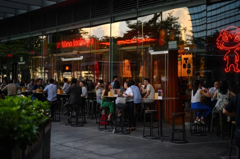 A wide, eye-level shot captures a lively outdoor dining scene at dusk, representing the file "1-Tono Izakaya Review.webp". Numerous patrons sit at high-top tables on a paved patio directly outside a modern restaurant with a large glass facade. Inside the building, bright red neon signs glow vividly, one spelling out "tōno izakaya" and another depicting a stylized, cartoonish monster character. The large glass windows reflect the warm hues of the setting sun and neighboring city buildings, while lush green bushes frame the bottom left corner, creating a bustling, urban nightlife atmosphere.