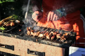 A close-up shot captures a chef actively grilling a long row of skewered meats on a traditional rectangular clay binchotan grill, representing the file "1-Yakitori at Home Enjoying Skewers Beyond the Grill.webp". Wisps of white smoke rise from the sizzling yakitori skewers as the chef, clad in a bright orange apron and wearing rings and wristbands, uses both hands to carefully turn the wooden sticks. To the left on the grill grate, fresh green vegetables and scallions are also being charred. The warm outdoor sunlight illuminates the textured surface of the grill and the cooking food, evoking a lively and appetizing culinary scene.