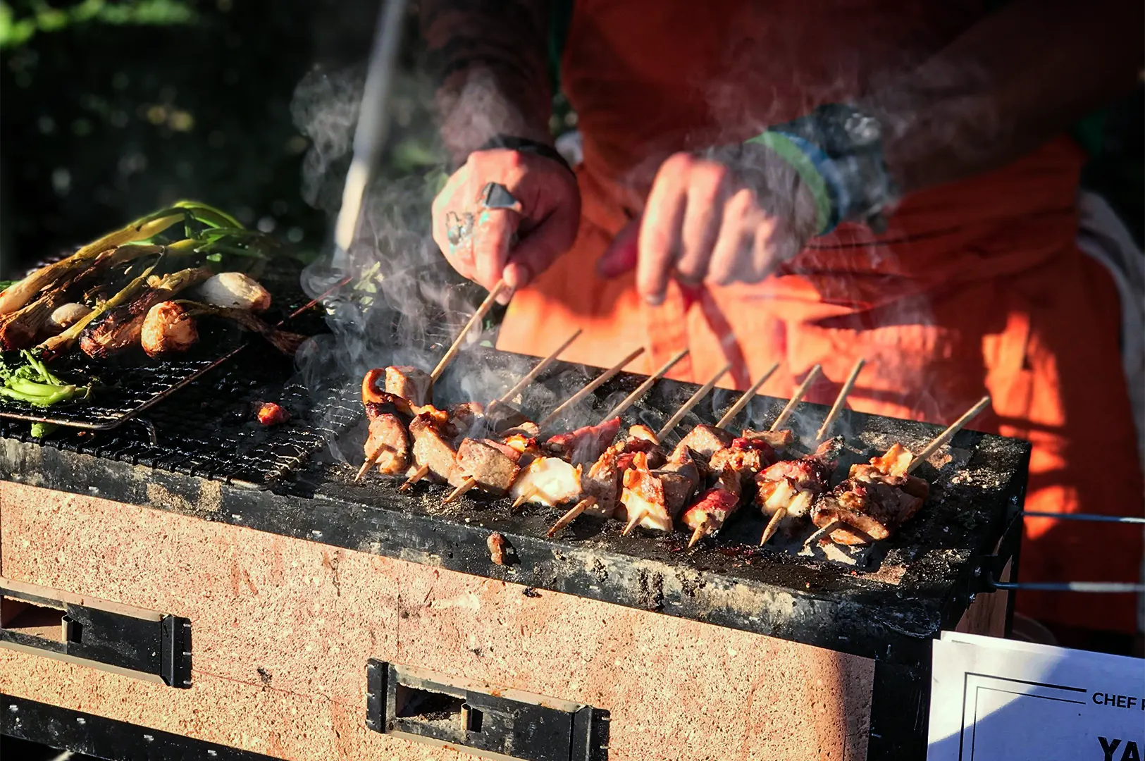 A close-up shot captures a chef actively grilling a long row of skewered meats on a traditional rectangular clay binchotan grill, representing the file "1-Yakitori at Home Enjoying Skewers Beyond the Grill.webp". Wisps of white smoke rise from the sizzling yakitori skewers as the chef, clad in a bright orange apron and wearing rings and wristbands, uses both hands to carefully turn the wooden sticks. To the left on the grill grate, fresh green vegetables and scallions are also being charred. The warm outdoor sunlight illuminates the textured surface of the grill and the cooking food, evoking a lively and appetizing culinary scene.