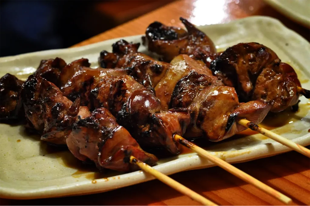 A high-angle, close-up shot captures a serving of hatsu (chicken heart) or reba (chicken liver) yakitori skewers beautifully presented on a platter, representing the file "11-Types of Yakitori Chicken Skewers in Singapore.webp". Four flat, paddle-style bamboo skewers are stacked on top of each other, holding plump, tender pieces of meat that are smothered in an exceptionally thick, dark, and glossy tare sauce that pools slightly onto the plate. The skewers rest on a wavy, textured white ceramic plate decorated with a few thin curls of green scallion on the side. In the background, a rustic wooden table holds a bowl of green edamame and a small dish containing Japanese spice blend, completing the izakaya-style setting.