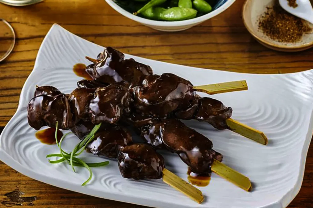 A high-angle, close-up shot captures a serving of hatsu (chicken heart) or reba (chicken liver) yakitori skewers beautifully presented on a platter, representing the file "11-Types of Yakitori Chicken Skewers in Singapore.webp". Four flat, paddle-style bamboo skewers are stacked on top of each other, holding plump, tender pieces of meat that are smothered in an exceptionally thick, dark, and glossy tare sauce that pools slightly onto the plate. The skewers rest on a wavy, textured white ceramic plate decorated with a few thin curls of green scallion on the side. In the background, a rustic wooden table holds a bowl of green edamame and a small dish containing Japanese spice blend, completing the izakaya-style setting.