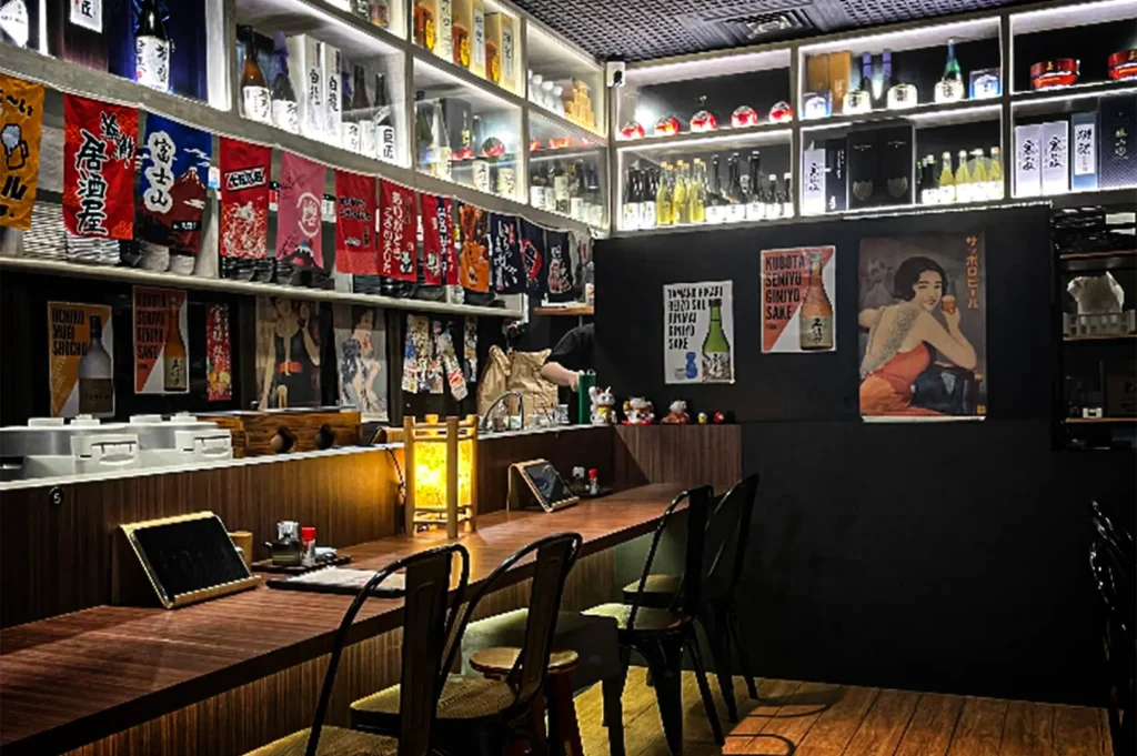 An eye-level, medium shot captures the cozy, atmospheric interior of a Japanese izakaya-style restaurant, representing the file "2-Best Fortune Centre Food for Japanese Dining.webp". The focus highlights a long, polished L-shaped wooden counter lined with dark metal chairs and a couple of wooden stools atop a hardwood floor. Each dining spot on the counter is equipped with a digital menu tablet, while a glowing traditional lantern and small condiment sets add to the intimate setup. The back wall is painted a dark charcoal color, adorned with vintage Japanese beverage posters and a few small Maneki-neko (lucky cat) figurines. Above the counter area, built-in, illuminated display shelves are densely stocked with various sake and liquor bottles, accented by a row of colorful, hanging Japanese noren curtains and flags that complete the authentic pub dining aesthetic.