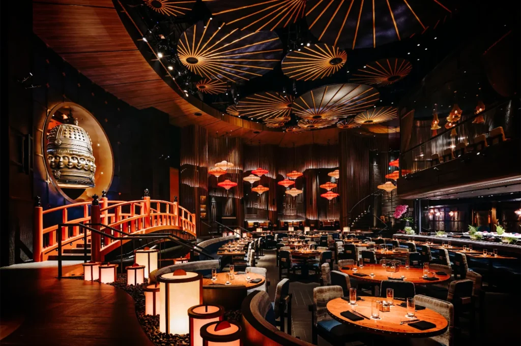 A wide, eye-level shot captures the dramatic, multi-tiered interior of a spacious Asian-inspired luxury restaurant, representing the file "2-Best Yakitori in Marina Bay Sands Food 6 Spots.webp". The main dining floor is filled with neatly arranged round and rectangular wooden tables surrounded by plush upholstered chairs, while glowing cylindrical lanterns line a curved walkway in the foreground. On the left, a prominent, brightly lit red wooden arch bridge spans over a pebble bed, leading toward a large circular cutout wall that showcases a massive, ornate bronze bell adorned with sculpted faces. The soaring, dark ceiling is decorated with massive, backlit circular umbrellas that radiate warm golden lines, complementing rows of hanging red and white paper lanterns in the background and creating a deeply atmospheric, theatrical dining space.