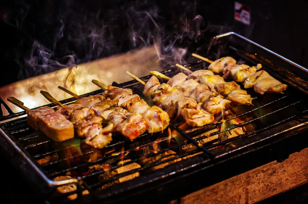 A close-up shot of several rows of chicken and pork skewers cooking over an open flame grill. The meat is sizzling on a metal grate, with visible grill marks and glazes catching the light. Wisps of heavy smoke curl upward from the glowing hot coals underneath, creating a warm, intense atmosphere.