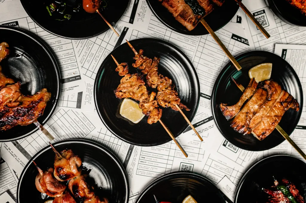 A top-down, flat-lay shot showcases a variety of Japanese yakitori and grilled skewers served in small black bowls, representing the file "2-Japanese Chicken Skewers Explained.webp". Positioned in the center, a black plate holds two crispy, golden-brown skewers—likely chicken skin or giblets—accompanied by a lemon wedge. Surrounding it are other small black dishes containing different grilled delicacies, including charred chicken wings on the left, juicy meat pieces on the bottom-left, thick-cut grilled pork belly or fish on the right, and skewers topped with green sauce at the top. The plates are arranged across a table surface fully covered with printed newspaper-style paper menus displaying grids, text, and QR codes, creating an organized, rustic, and appetizing culinary layout.