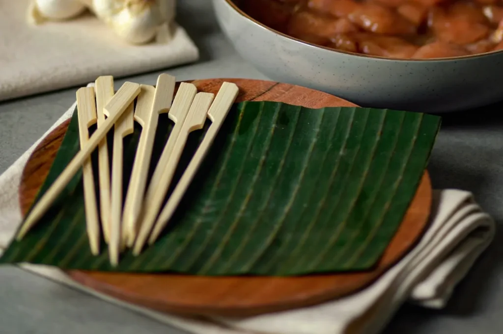 A close-up, high-angle shot captures a collection of paddle-style bamboo skewers arranged on a round wooden platter, representing the file "2-Not Just a Stick The Role of Bamboo.webp". The flat-handled skewers are piled together on the left side of a fresh, green banana leaf that serves as a decorative liner over the warm-toned wooden plate, which rests on a folded white cloth. In the softly blurred background, a grey ceramic bowl holds marinated chunks of raw meat, likely chicken, alongside a bulb of garlic, setting the stage for food preparation against a dark, textured tabletop.