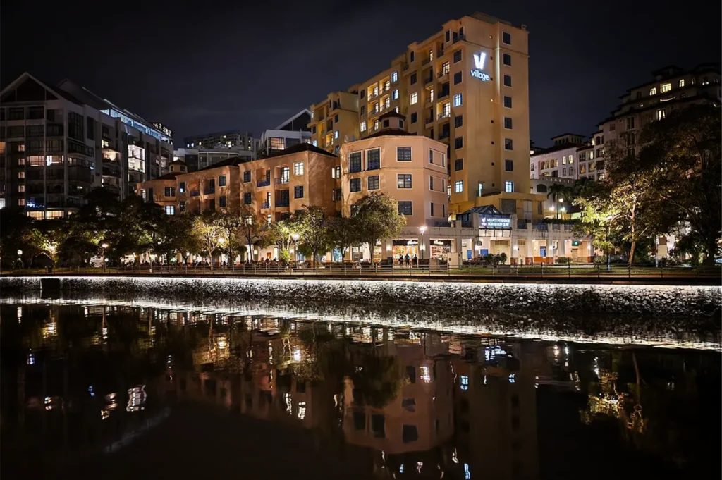 A night shot captures a row of illuminated, warm-toned apartment and hotel buildings, including the "Village Residence Robertson Quay," lined up along a waterfront. The structures feature a mix of beige and orange facades with glowing windows, which cast a vibrant, shimmering reflection onto the dark, still surface of the river below. A stone embankment separates the water from a paved walkway lined with lush green trees and streetlights, while a dark, overcast night sky provides a stark contrast to the brightly lit urban scene.