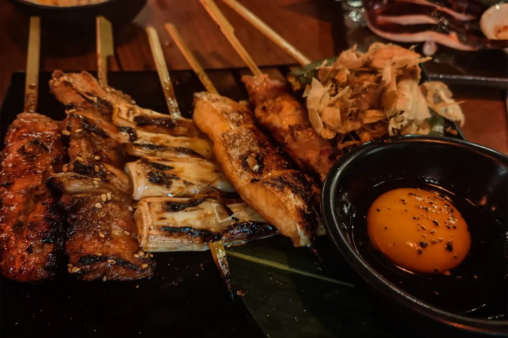 A high-angle, close-up shot captures a delectable assortment of Japanese yakitori skewers presented on a black rectangular plate over a green leaf, representing the file "2-TORIMARO Singapore Review.webp". Arranged tightly from left to right are various grilled skewers, including glistening, caramelized minced meat tsukune sprinkled with white sesame seeds, charred white scallions or leeks, a juicy piece of salmon or pork belly, and tender chicken chunks. In the upper right corner of the plate sits a side portion of grilled vegetables covered in a mound of thin, translucent bonito flakes. Dominating the lower right foreground is a small, round black dipping bowl containing a bright, glossy raw egg yolk seasoned with cracked black pepper, all illuminated by warm, dim restaurant lighting that accentuates the rich textures of the izakaya-style meal.